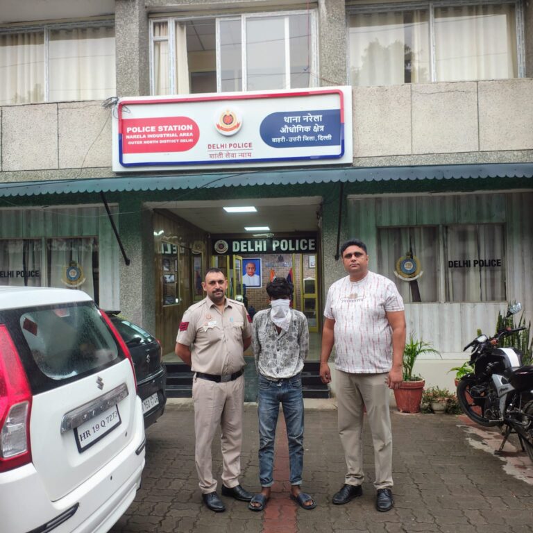 नरेला इंडस्ट्रियल एरिया थाने के बाहर दो लोगों के साथ चेहरे पर कपड़ा बांधे एक व्यक्ति, जिनमें एक वर्दीधारी है।